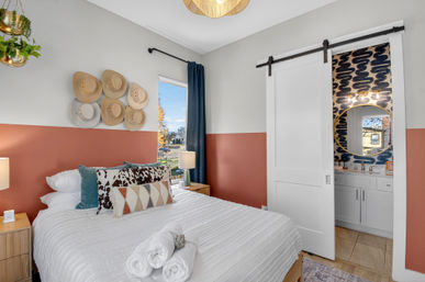 Sunlit modern bedroom with white bedding, patterned throw pillows and rolled towels, terracotta half-painted walls accented by decorative straw hats, navy curtain, wooden nightstands and a sliding barn-door to a bathroom with a round mirror and bold blue wallpaper.