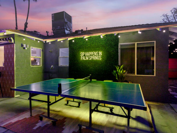 Dusk Palm Springs patio with a ping-pong table under string lights, neon sign reading 'SIP HAPPENS IN PALM SPRINGS', and silhouetted palm trees.