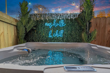 Bubbling backyard hot tub at dusk with neon "Nashville & chill" sign on a greenery wall, string lights and tropical plants
