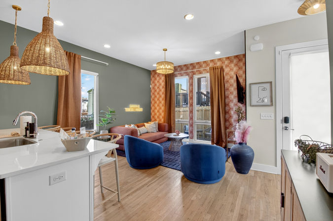 Open-concept modern living room and kitchen with white island and sink, wicker pendant lights, blue velvet swivel chairs, rust-colored sofa, geometric orange accent wallpaper, wood floors, indoor plant and glass patio doors letting in natural light.