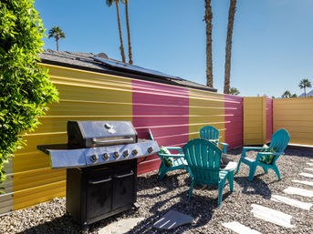 Sunny desert-style backyard patio with tall palm trees, colorful yellow-and-pink corrugated fence, black gas grill, four turquoise Adirondack chairs around a small fire pit on gravel with stepping stones and solar panels on a nearby roof.