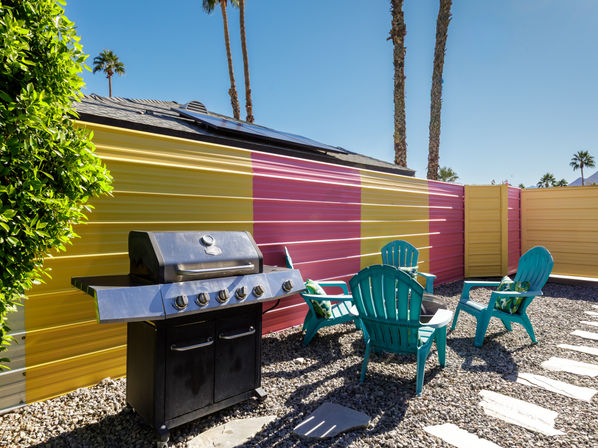 Sunny desert-style backyard patio with tall palm trees, colorful yellow-and-pink corrugated fence, black gas grill, four turquoise Adirondack chairs around a small fire pit on gravel with stepping stones and solar panels on a nearby roof.