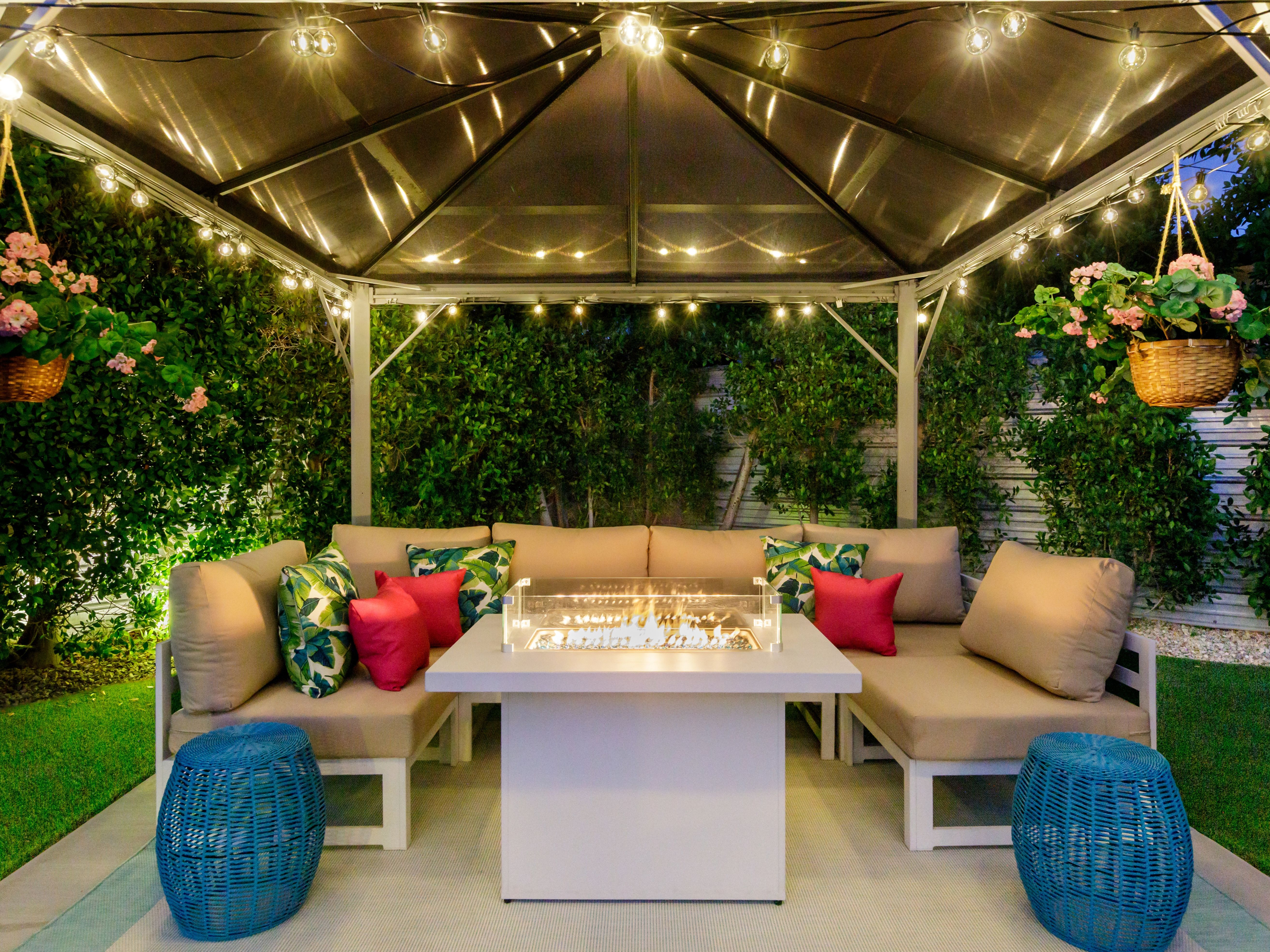 Cozy backyard gazebo lounge in the evening with cushioned L-shaped sofa, tropical-print and pink pillows, central glass-enclosed fire pit table, string lights overhead, hanging flower baskets, blue wicker stools and lush hedges.