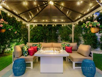 Cozy backyard gazebo lounge in the evening with cushioned L-shaped sofa, tropical-print and pink pillows, central glass-enclosed fire pit table, string lights overhead, hanging flower baskets, blue wicker stools and lush hedges.