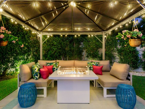 Cozy backyard gazebo lounge in the evening with cushioned L-shaped sofa, tropical-print and pink pillows, central glass-enclosed fire pit table, string lights overhead, hanging flower baskets, blue wicker stools and lush hedges.