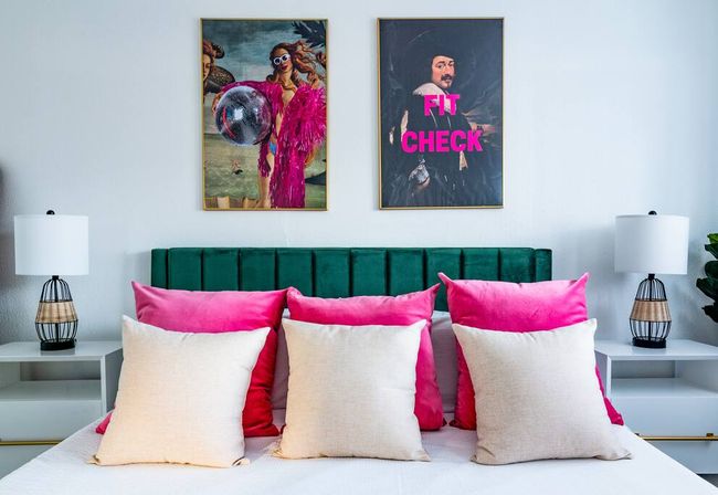 Stylish modern bedroom with emerald velvet headboard, three hot-pink accent pillows and three beige throw pillows on a white bed, two framed pop-art prints above (one reads “FIT CHECK”) and matching bedside lamps.