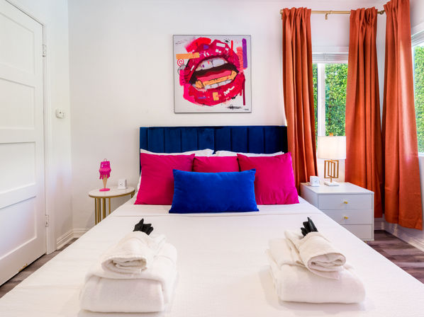 Sunlit modern bedroom with navy velvet headboard, white bed dressed with bright blue and hot-pink pillows, neatly folded white towels at the foot, orange curtains, bedside lamps, and a bold pop-art lips painting above the bed.