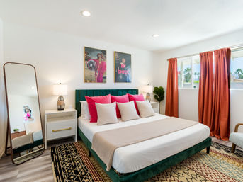 Sunlit modern bedroom with emerald green velvet bed, bright pink accent pillows, full-length mirror, patterned rugs, coral curtains, bedside lamps and a window view of palm trees.