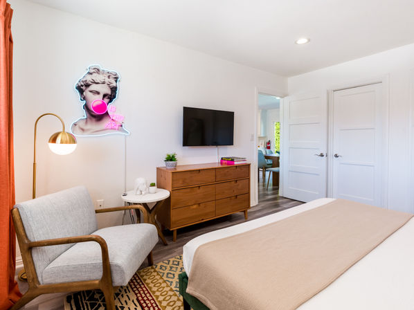 Bright modern bedroom interior with a mid-century wooden dresser and wall TV, gray armchair and brass floor lamp, bed with beige throw, patterned rug, and a neon pop-art bust blowing bubble gum on the wall.