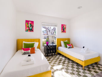 Sunlit guest bedroom with two yellow upholstered twin beds, white linens and folded towels, colorful cactus wall art, green and pink accent pillows, black geometric nightstand under a window with a tree view, and a green-and-white checkered rug.