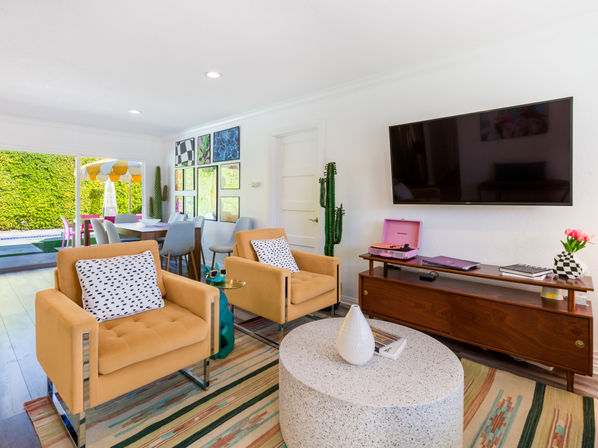 Sunny modern living room in a suburban home with two mustard armchairs, terrazzo coffee table, wall-mounted TV, pink record player on a wood console, colorful rug and dining area opening to a backyard pool with a yellow-striped umbrella.