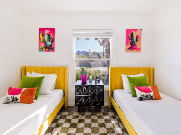 Sunlit twin bedroom with yellow velvet headboards, vibrant cactus artwork, green and pink accent pillows, black geometric nightstand with potted cactus, and a window framing a desert landscape with distant mountains.