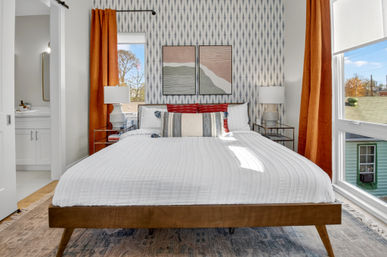 Bright mid-century modern bedroom with wooden platform bed, crisp white quilt, striped and red accent pillows, geometric wallpaper, orange curtains, glass bedside tables with lamps, abstract art above bed and a large window with neighborhood view.