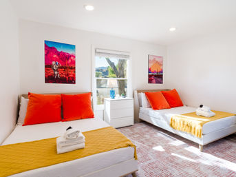 Sunlit modern guest bedroom with two white beds, bold orange pillows and mustard throws, white dresser with blue lamp, window showing a tree and hills, and vibrant desert-themed wall art.