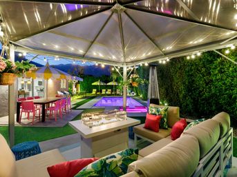 Evening backyard patio under a clear canopy with string lights — cushioned lounge seating with tropical pillows around a modern fire table, colorful outdoor dining with pink chairs and yellow-striped umbrellas, illuminated rectangular pool and manicured hedge.