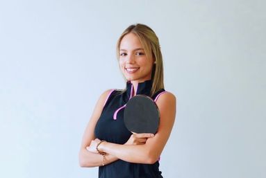 Smiling woman in a black athletic top holding a ping pong paddle with arms crossed against a bright indoor wall
