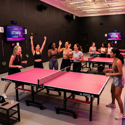 Group of women cheering and laughing around bright pink ping-pong tables in a modern indoor table tennis studio, holding paddles and drinks during a social ping-pong event.