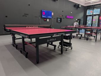 Modern indoor ping-pong room with bright pink table tennis tables, nets, rolling carts and digital booking screens in a sleek recreational space.