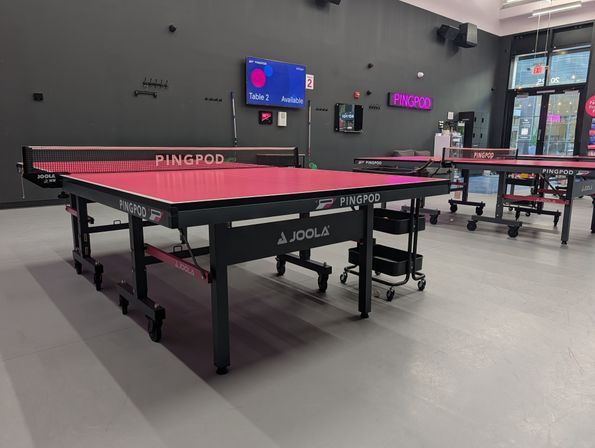 Modern indoor ping-pong room with bright pink table tennis tables, nets, rolling carts and digital booking screens in a sleek recreational space.
