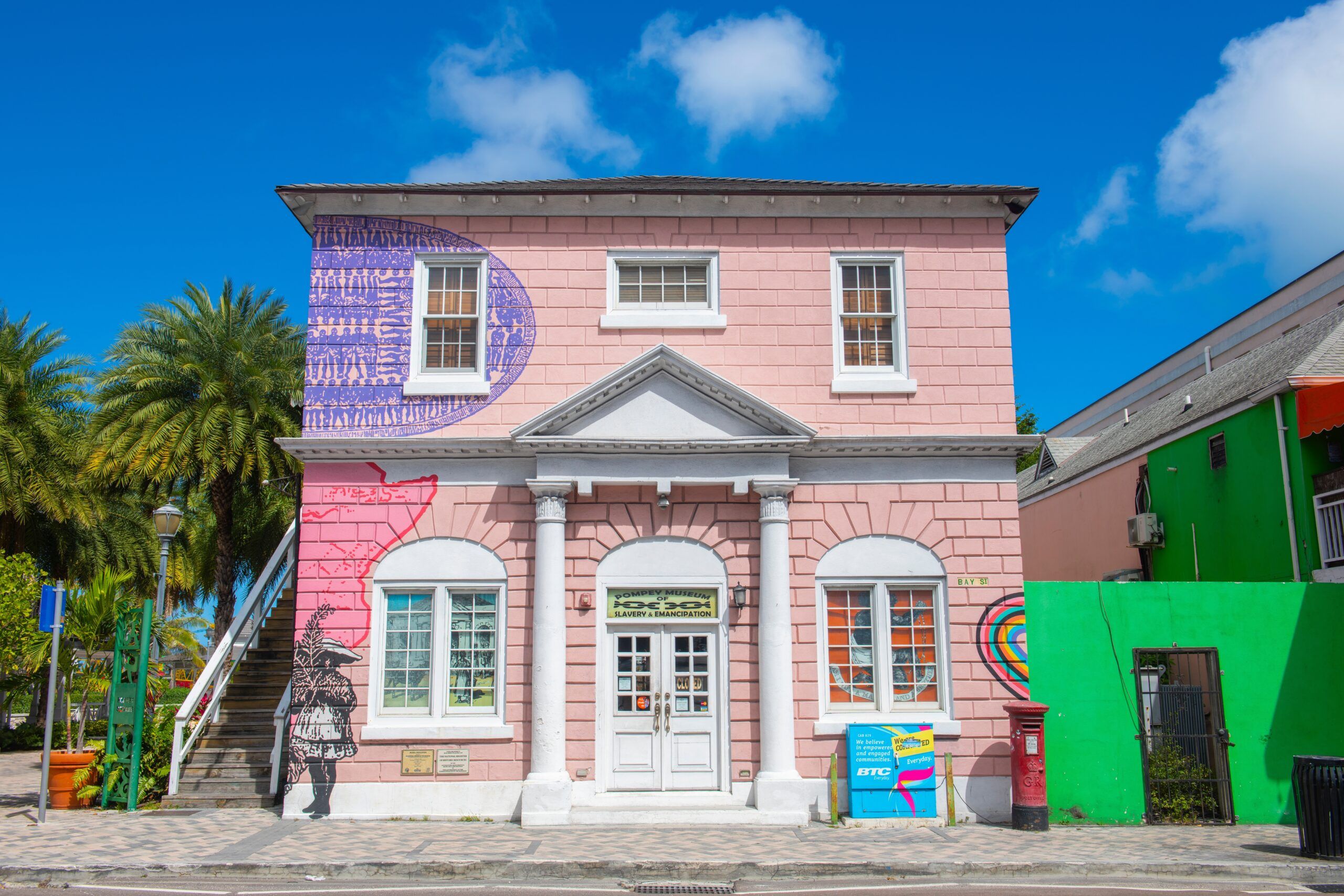 Pink colonial-style building with white columns and colorful murals, palm trees and bright blue sky on a sunny Caribbean street in Nassau, Bahamas