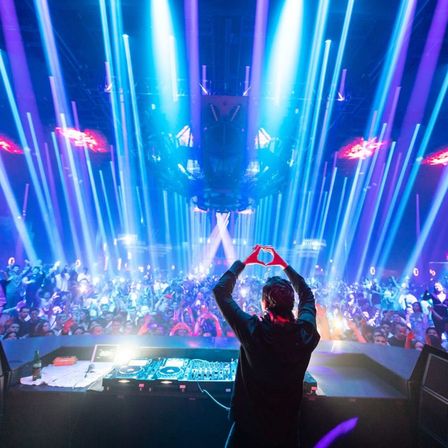 Nightclub DJ forming a heart with hands over a packed dance floor, blue and purple laser beams and an energetic EDM crowd