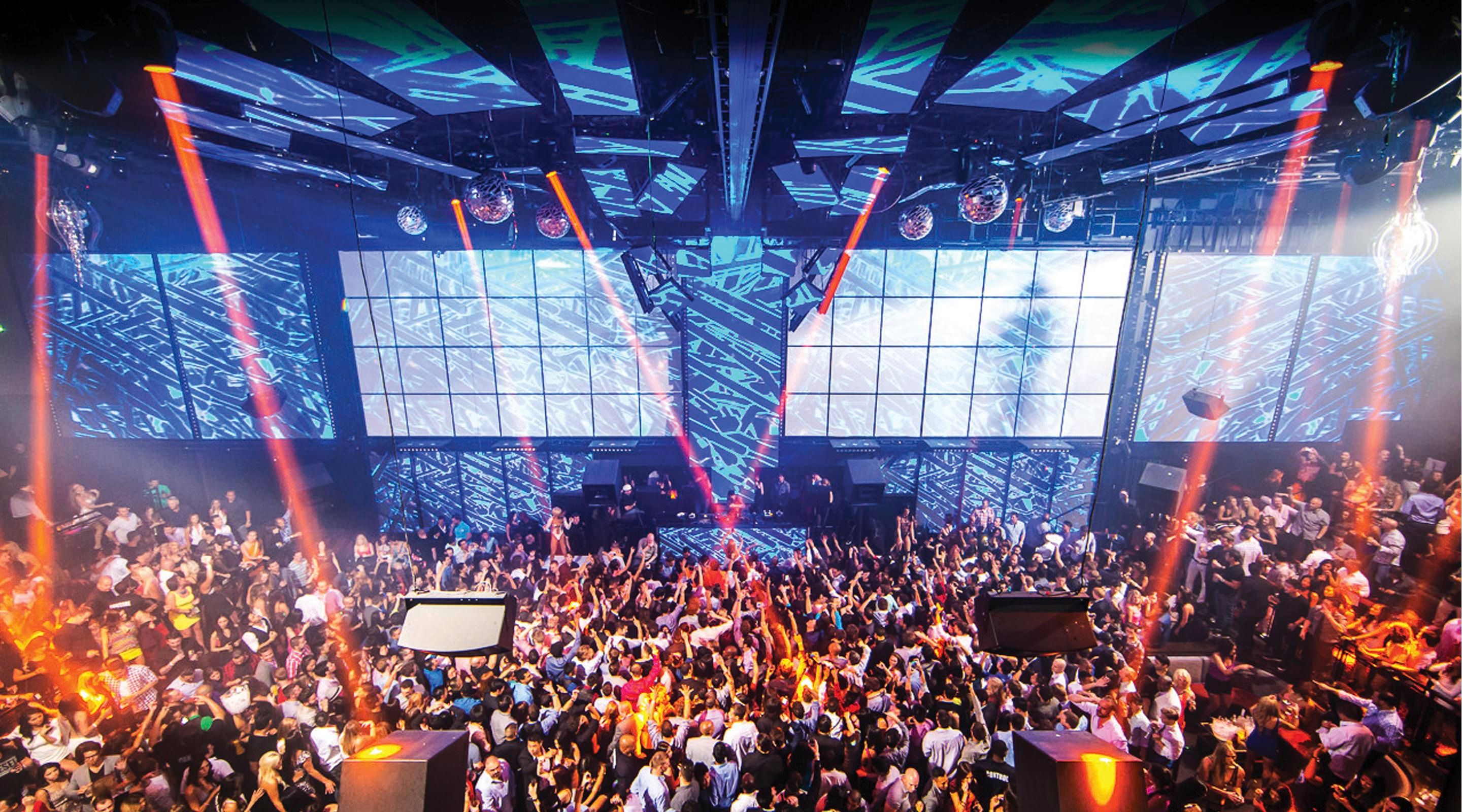 Packed indoor nightclub dance floor with a DJ stage, oversized LED video wall, disco balls and colorful laser beams lighting a cheering crowd