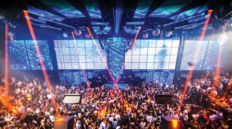 Packed indoor nightclub dance floor with a DJ stage, oversized LED video wall, disco balls and colorful laser beams lighting a cheering crowd
