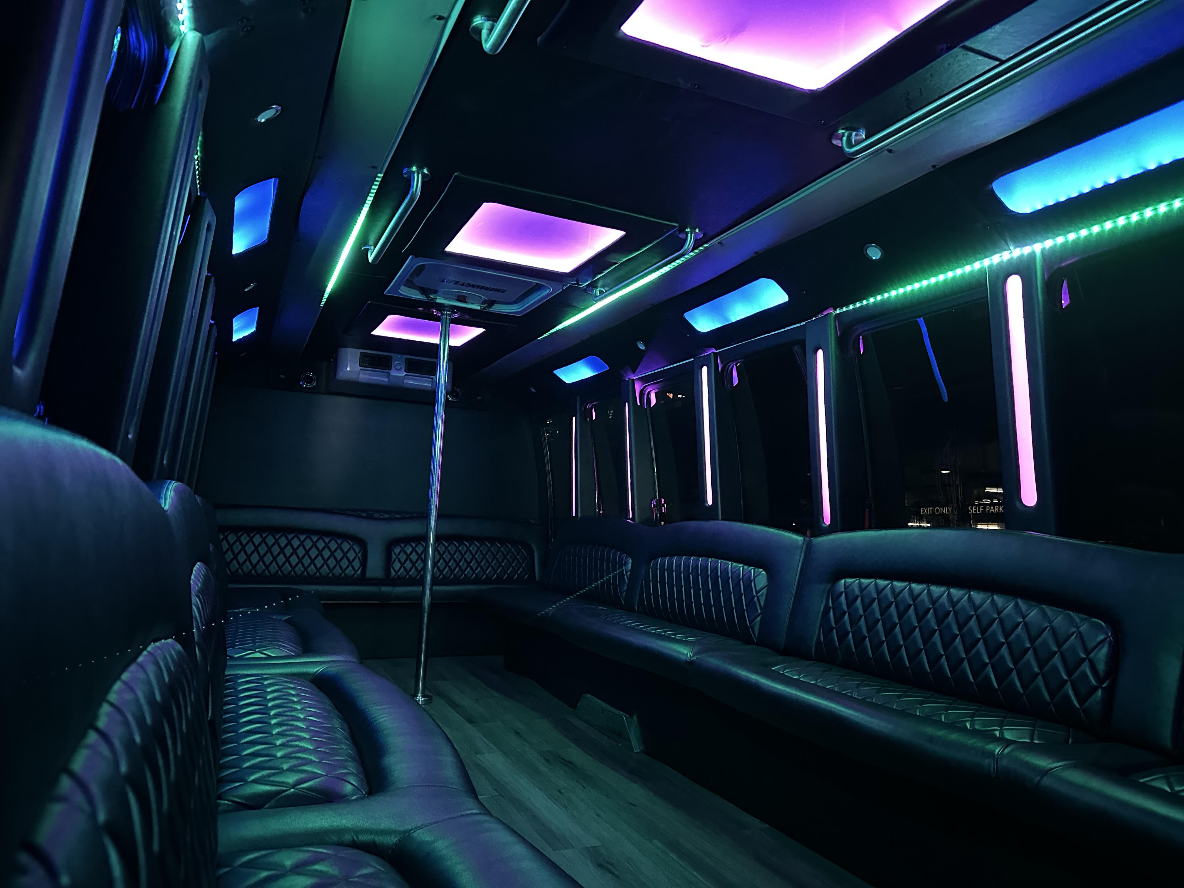 Party bus interior with quilted black leather bench seating, central dance pole and vibrant purple, teal and blue LED ceiling and strip lights — nightclub-style mobile lounge.