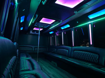 Party bus interior with quilted black leather bench seating, central dance pole and vibrant purple, teal and blue LED ceiling and strip lights — nightclub-style mobile lounge.