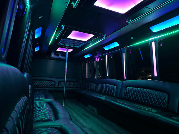 Party bus interior with quilted black leather bench seating, central dance pole and vibrant purple, teal and blue LED ceiling and strip lights — nightclub-style mobile lounge.