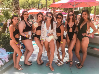 Seven women in swimsuits posing at a sunny resort pool party with pink umbrellas and palm trees, smiling and holding canned drinks and a coconut