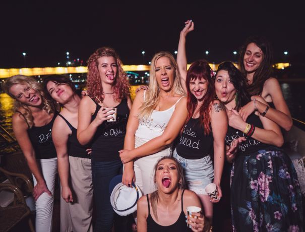 Bachelorette party at a riverside dock at night — a laughing group of women in matching tanks and one in white, holding drinks with city lights and a lit bridge behind them