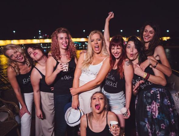 Bachelorette party at a riverside dock at night — a laughing group of women in matching tanks and one in white, holding drinks with city lights and a lit bridge behind them