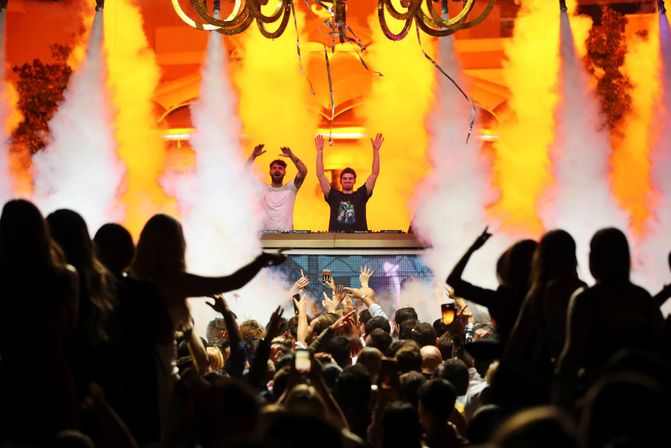 Packed nightclub dancefloor with two DJs on a raised stage, orange lights and smoke jets, crowd cheering with hands and phones aloft