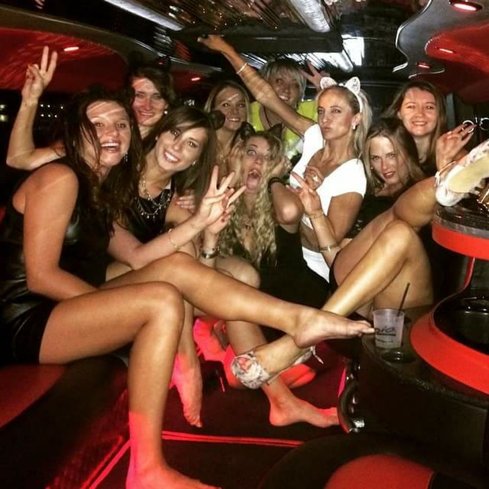 Group of women laughing and posing inside a red-lit stretch limousine on a city night out, wearing party dresses, cat-ear headband, some barefoot and holding drinks — lively bachelorette or girls' night celebration.