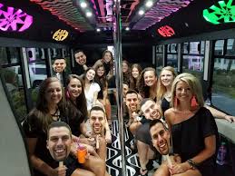 Smiling group of friends on a brightly lit party bus interior with neon lights, a central dance pole and drinks, celebrating a night out.