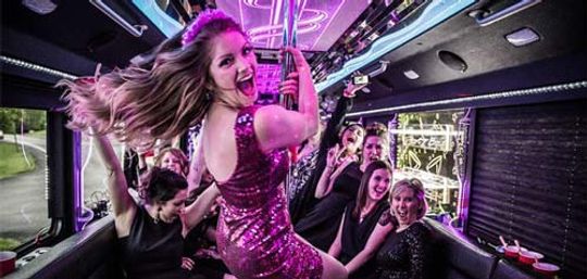Women celebrating a bachelorette-style party inside a neon-lit party bus, a woman in a pink sequin dress dancing on a pole while friends cheer and laugh.