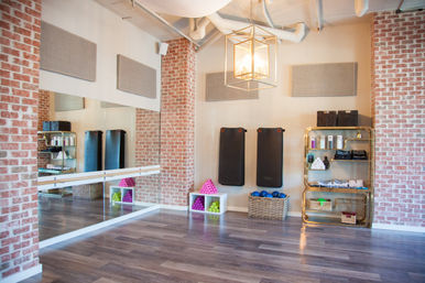 Bright boutique fitness studio interior with exposed brick, mirrored wall and ballet barre, three black workout mats, colorful hand weights and blue exercise balls in baskets, gold shelving with towels and props, wood-look floor