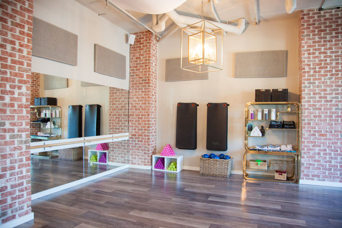 Bright boutique fitness studio interior with exposed brick, mirrored wall and ballet barre, three black workout mats, colorful hand weights and blue exercise balls in baskets, gold shelving with towels and props, wood-look floor