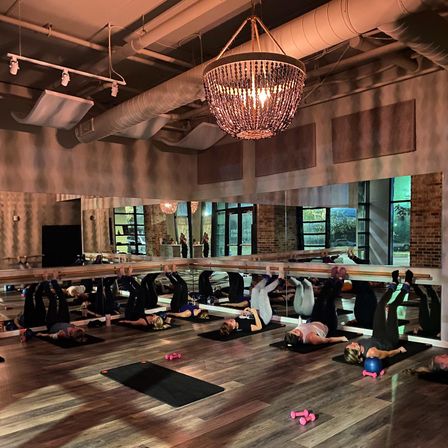 Boutique barre workout in a mirrored fitness studio with participants on mats pressing feet to the barre, pink dumbbells and a small blue exercise ball on wood floors, a beaded chandelier and exposed ductwork overhead.