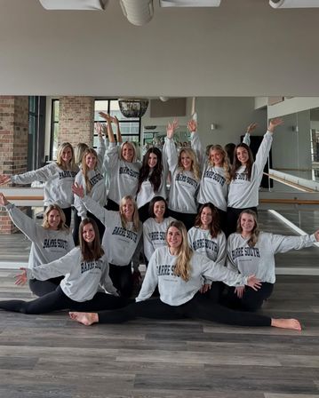 Cheerful group of women in a barre/dance studio wearing matching gray sweatshirts and black leggings, posing in front of mirrors and exposed brick with two dancers doing full splits on the wood floor and others raising their arms.