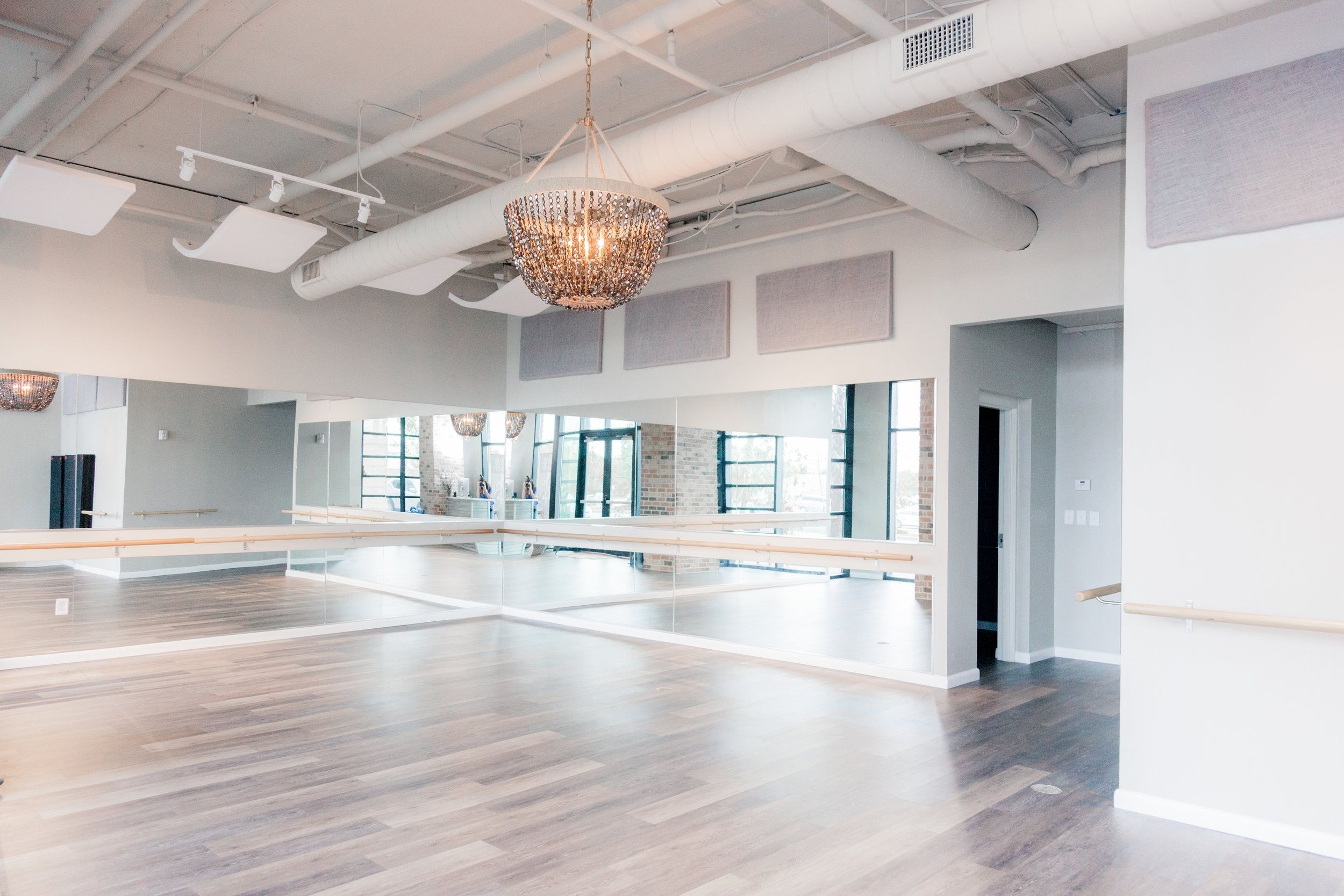 Bright modern dance studio with light wood floors, wall-length mirrors, ballet barres, a sparkling chandelier, exposed industrial ceiling and large windows