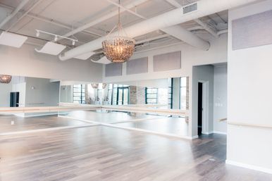 Bright modern dance studio with light wood floors, wall-length mirrors, ballet barres, a sparkling chandelier, exposed industrial ceiling and large windows