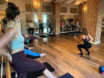 Energetic barre-style fitness class in a mirrored brick-walled studio, instructor squatting at front while participants balance on mats and a ballet barre on hardwood floors.
