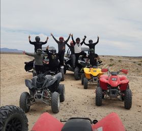 Group of helmeted riders cheering on ATVs and a side-by-side UTV parked on a sandy desert trail — off-road ATV/UTV adventure in open desert under a cloudy sky.