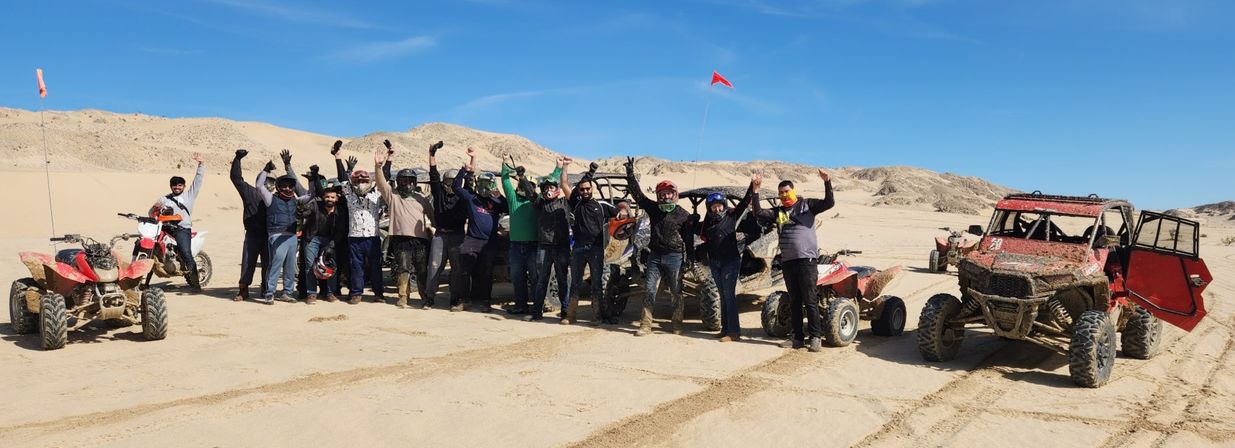 Off-road riders raising their arms while posing with muddy ATVs and a red UTV on sunlit desert sand dunes under a clear blue sky
