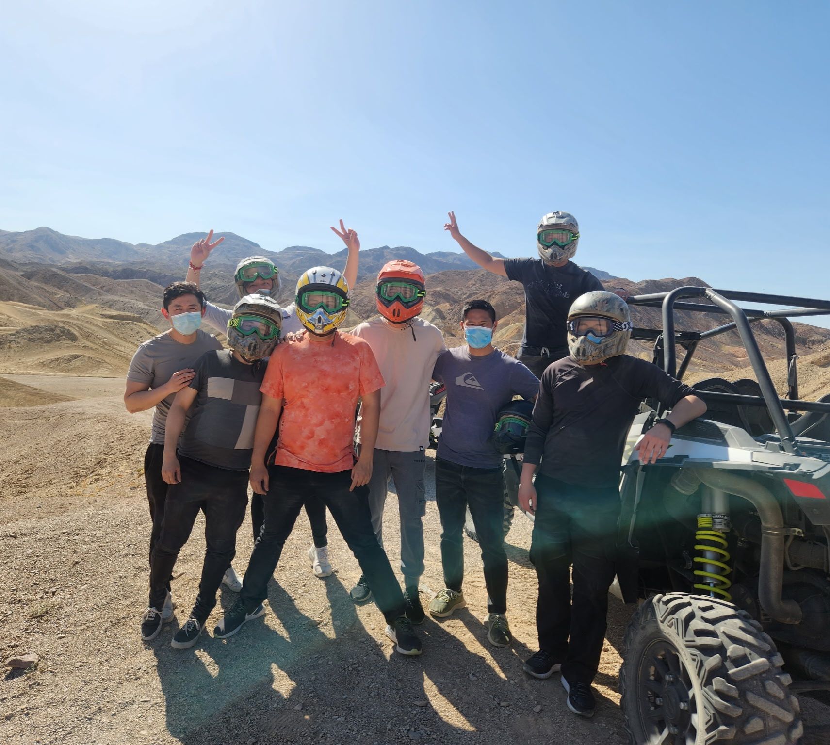 Group of friends in helmets, goggles and face masks posing by a UTV on sunny desert dunes for an off‑road ATV/UTV adventure with rocky mountain backdrop