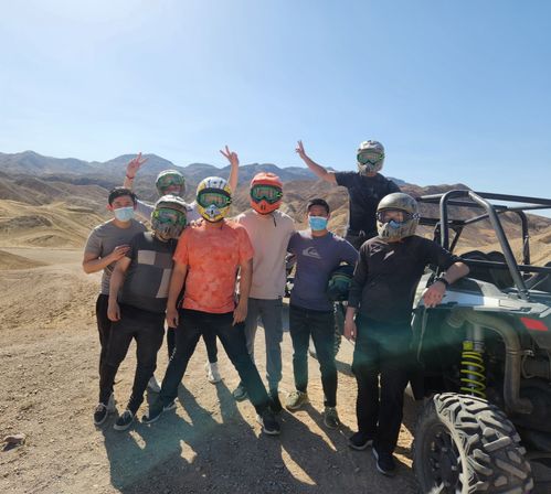 Group of friends in helmets, goggles and face masks posing by a UTV on sunny desert dunes for an off‑road ATV/UTV adventure with rocky mountain backdrop