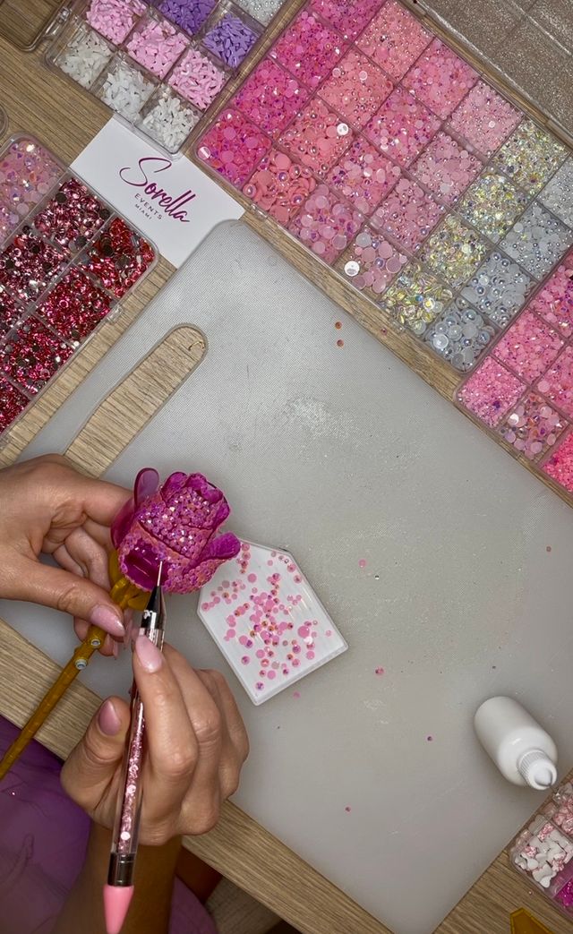 Close-up of hands using a rhinestone picker to attach pink sequins and rhinestones to a decorative flower on a DIY craft workspace, surrounded by organized pink and purple sequin trays and a glue bottle.