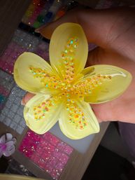 Hand holding a yellow resin flower decorated with iridescent sequins over a sparkly craft supply workstation